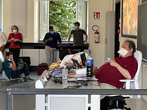 L'immagine mostra un gruppo di persone in un ambiente interno, probabilmente una sala conferenze o un laboratorio. Al centro, un uomo in maglietta rossa e pantaloni beige è seduto in una sedia bianca, con un'espressione rilassata e indossa una mascherina. Sul tavolo davanti a lui ci sono vari oggetti, tra cui una bottiglia di Pepsi, un computer portatile, e alcuni fogli. Sullo sfondo, altre persone sono in piedi o sedute, alcune con mascherine, e sembrano essere impegnate in attività diverse. Una finestra aperta lascia entrare luce naturale e si vede un paesaggio verde all'esterno. Sul muro c'è un'etichetta con la scritta "ESTINTORE" e un estintore rosso. L'atmosfera sembra informale e lavorativa.